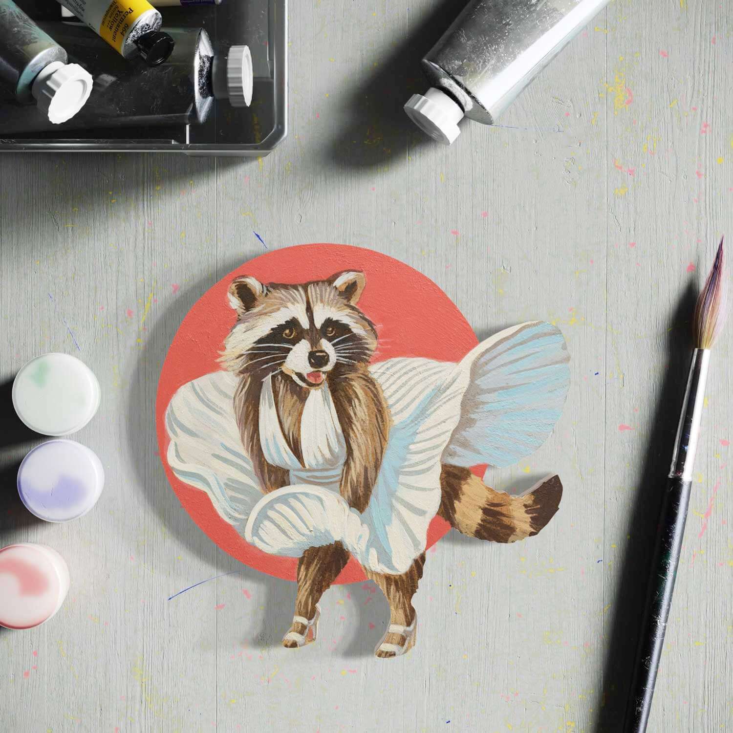 Hand-painted raccoon in a robe, circular art piece on a colorful workspace with paint supplies.